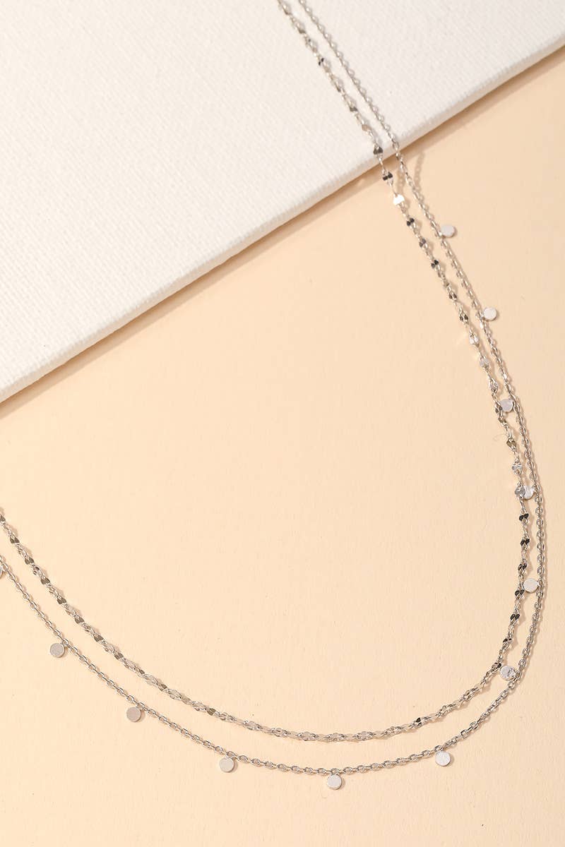 Delicate layered silver necklace with small disc accents styled on a beige background.