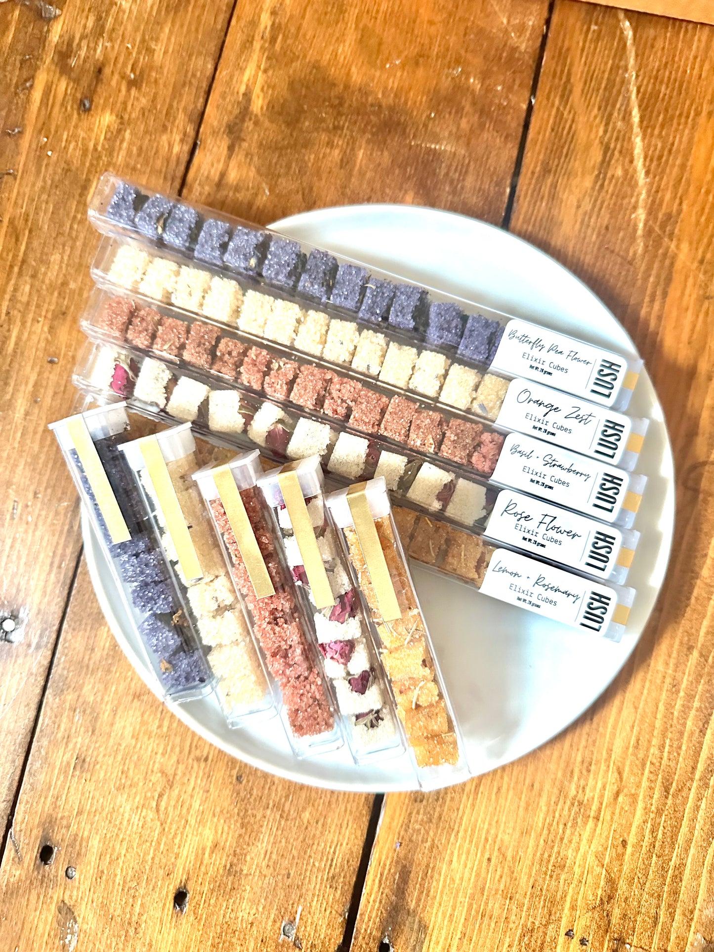 The Goodery Lush Elixir drink cubes in clear rectangular tube packaging, arranged on a white plate, with assorted flavors including butterfly pea flower, orange zest, rose flower, and lemon rosemary.