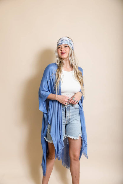 Woman wearing a blue kimono-style cardigan, white top, denim shorts, and floral headband against a beige background