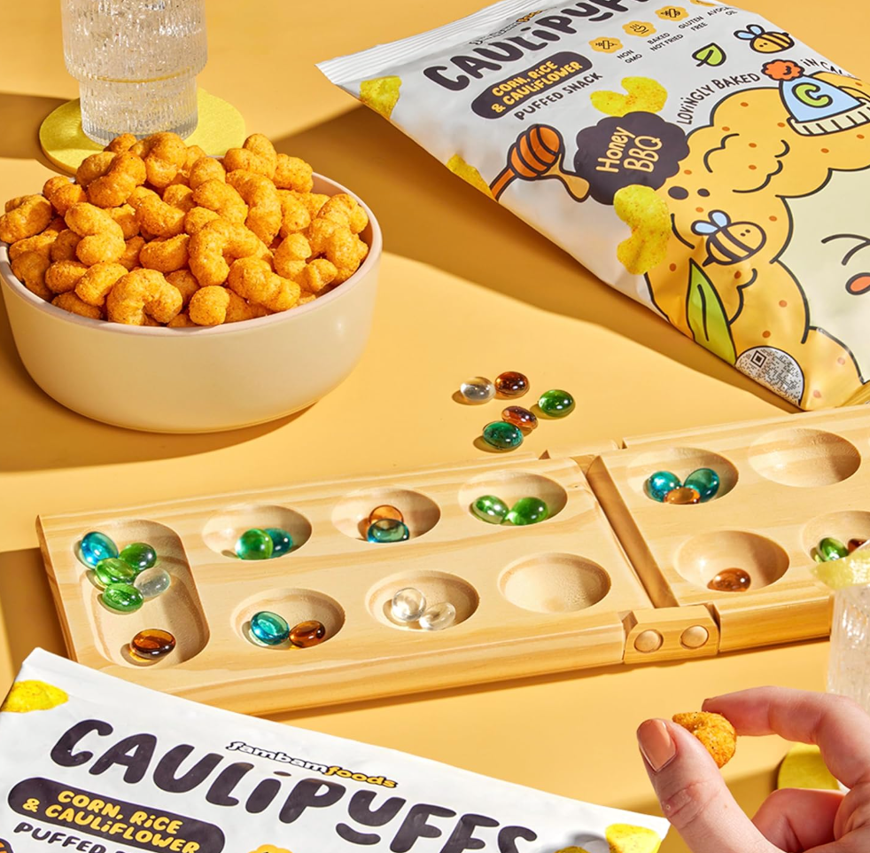 Bowl of Caulipuffs Honey BBQ puffed snacks with packaging, marbles, and a game board on a yellow background.