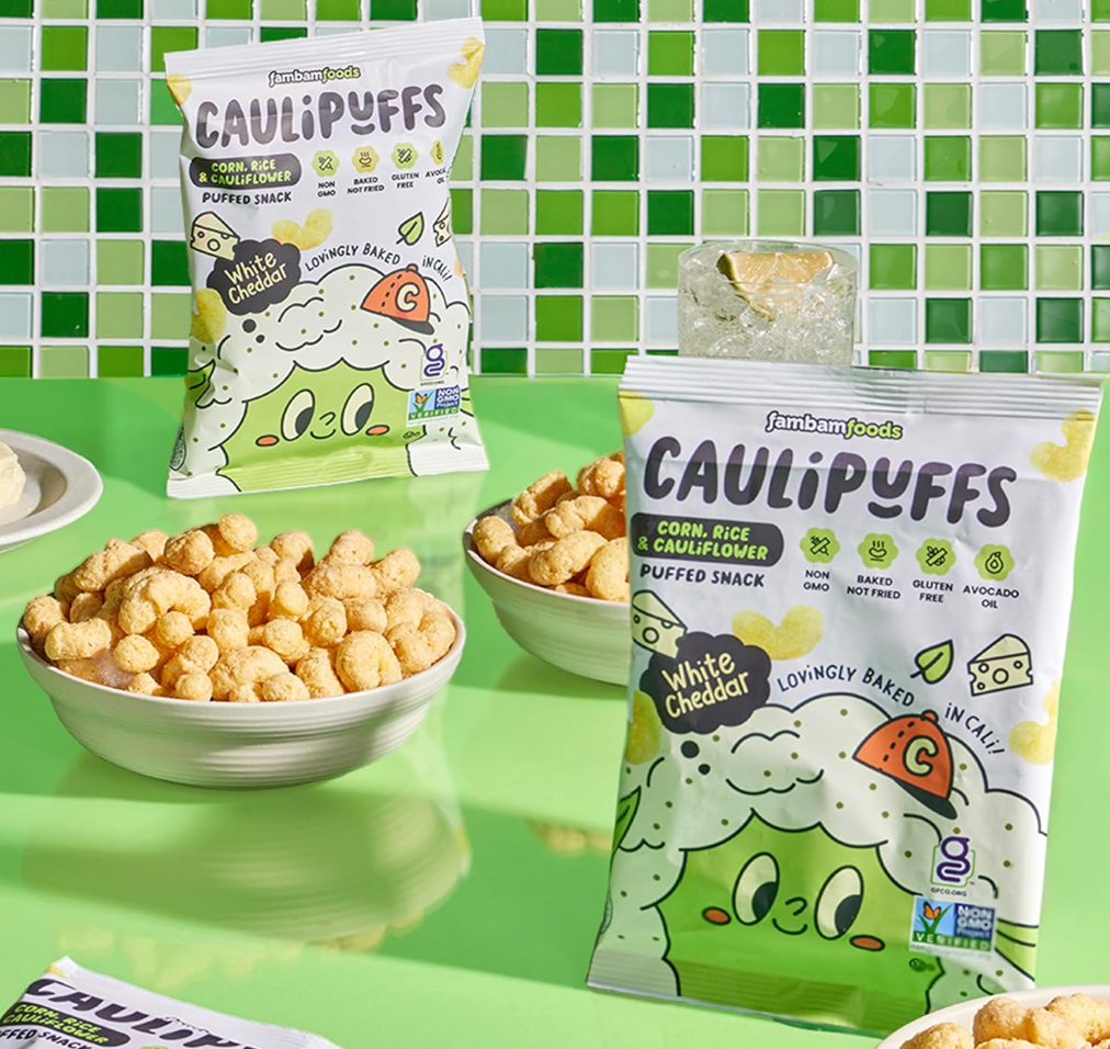 Bowls of Caulipuffs White Cheddar puffed snacks with packaging shown on a green table and checkered green-and-white tile background.