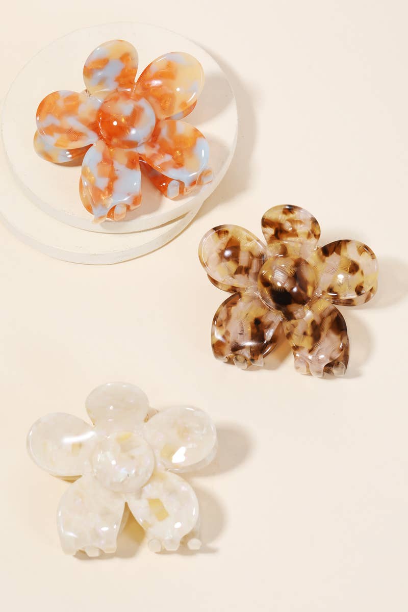 Set of three floral-shaped acetate hair clips in marbled orange, tortoiseshell brown, and pearly white on a beige background.