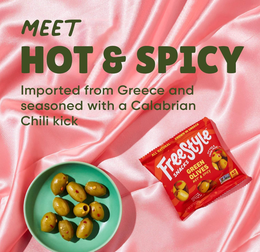 Freestyle Snacks Hot & Spicy Green Olives package with bowl of olives on a pink silk background, featuring text about Greek origin and Calabrian chili flavor.