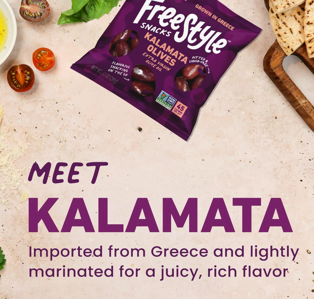 Freestyle Snacks Kalamata Olives package on a textured surface with tomatoes, pita bread, and olive oil, highlighting Greek origin and rich flavor.