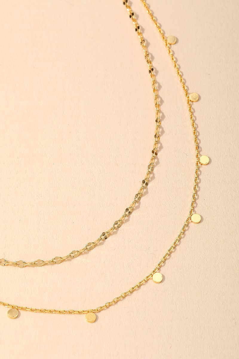 Close-up of two delicate gold layered necklaces with textured chain and small round pendants on a beige background.