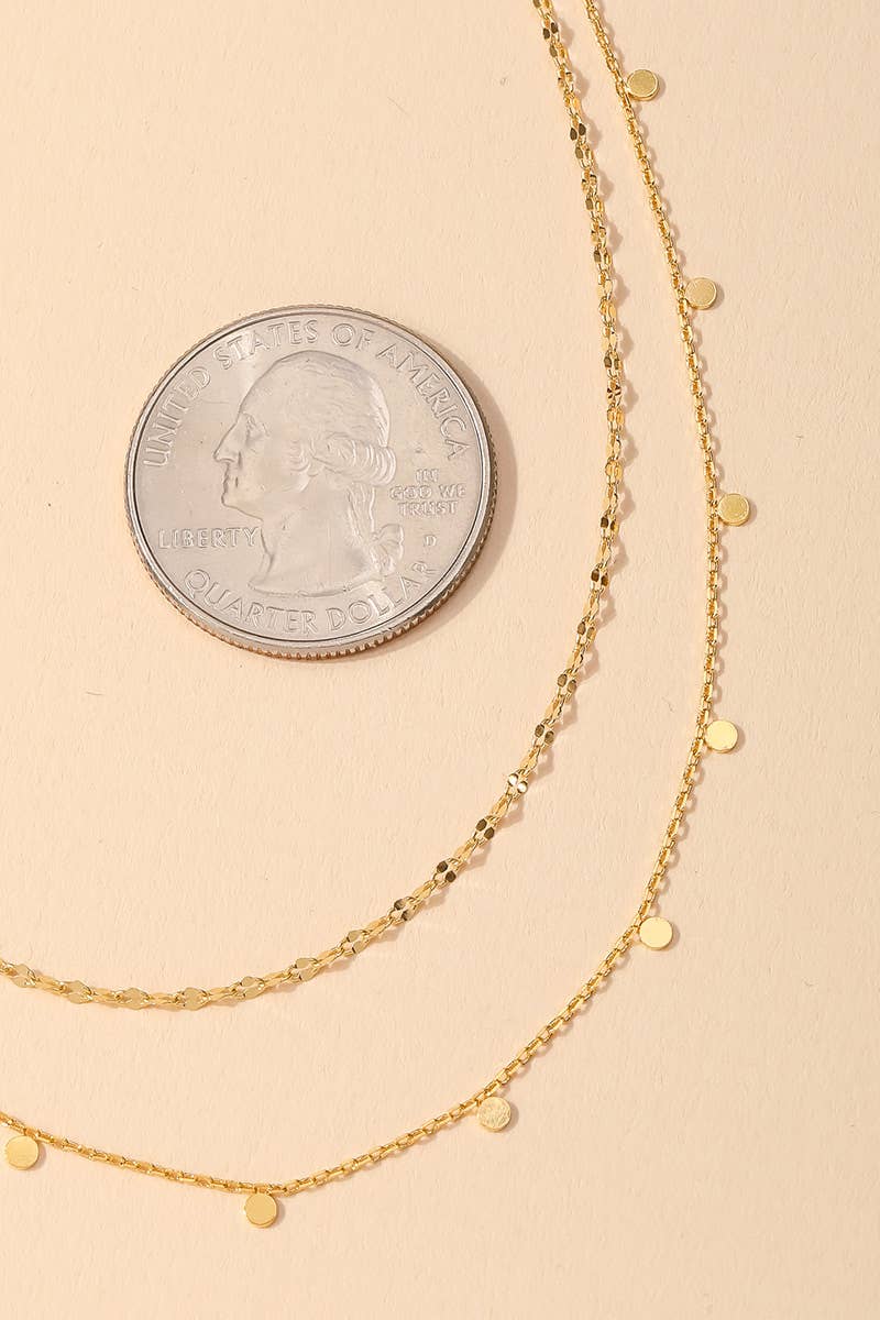 Two delicate gold layered necklaces with small round pendants shown beside a U.S. quarter for size reference on a beige background.