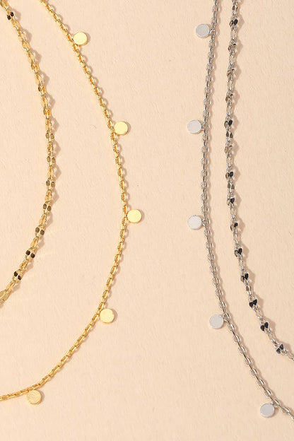 Gold and silver layered necklaces with delicate round pendants on a beige background.