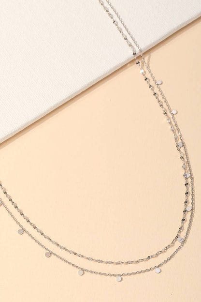 Delicate layered silver necklace with small disc accents styled on a beige background.