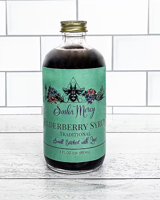 Sailor Mercy Elderberry Syrup in an 8 oz glass bottle with teal label, displayed on a white wooden surface with tiled wall background, small batched with love.