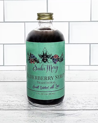 Sailor Mercy Elderberry Syrup in an 8 oz glass bottle with teal label, displayed on a white wooden surface with tiled wall background, small batched with love.