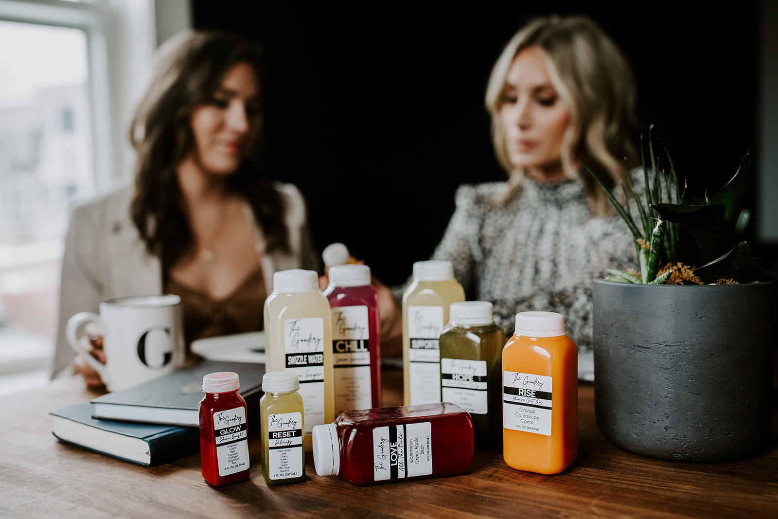 The Goodery cold-pressed juice bottles including Glow, Reset, Rise, and Chill displayed on a table with founders in background