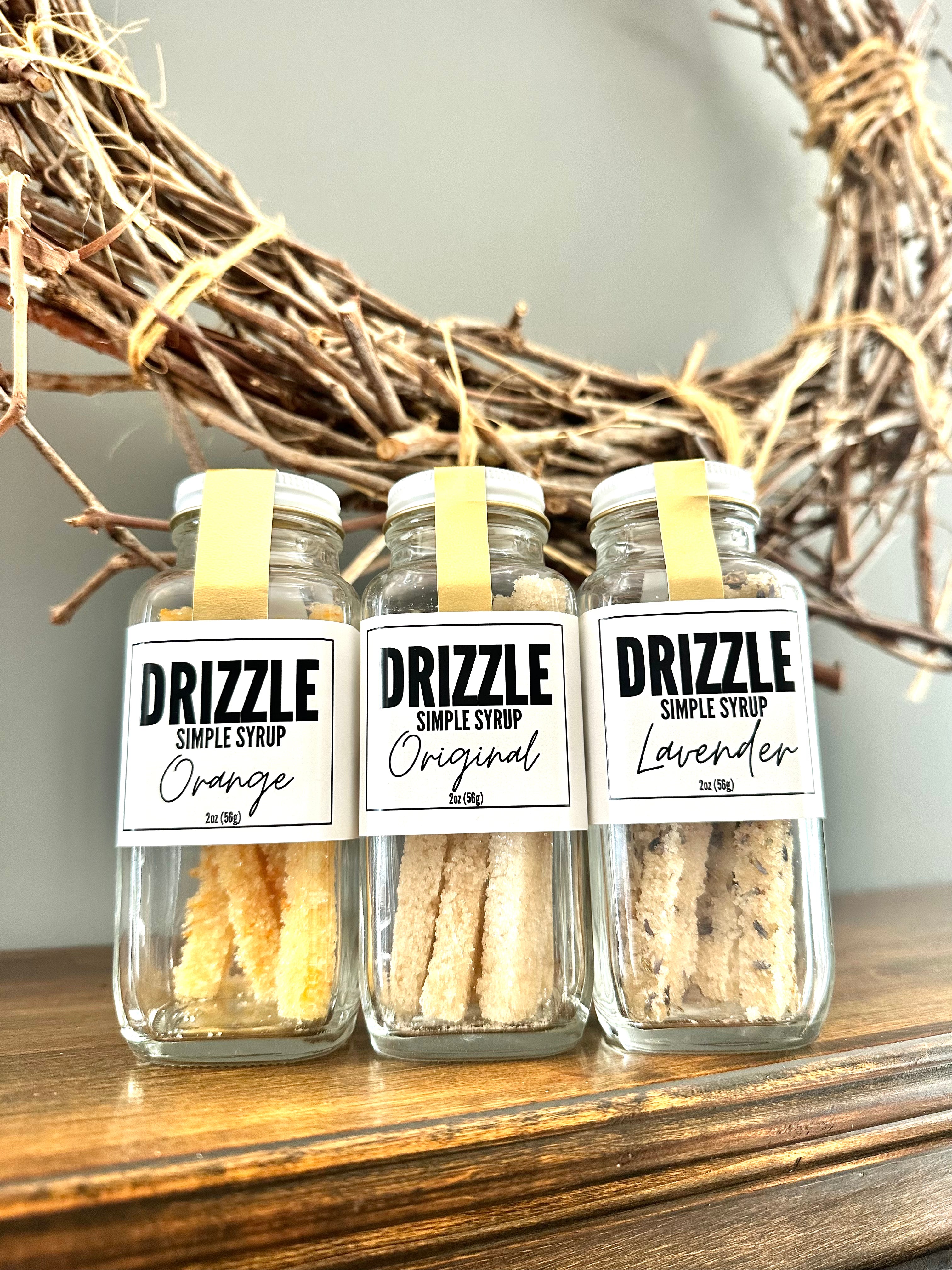The Goodery Drizzle Simple Syrup jars in flavors Orange, Original, and Lavender displayed on a wooden shelf with a rustic wreath in the background.