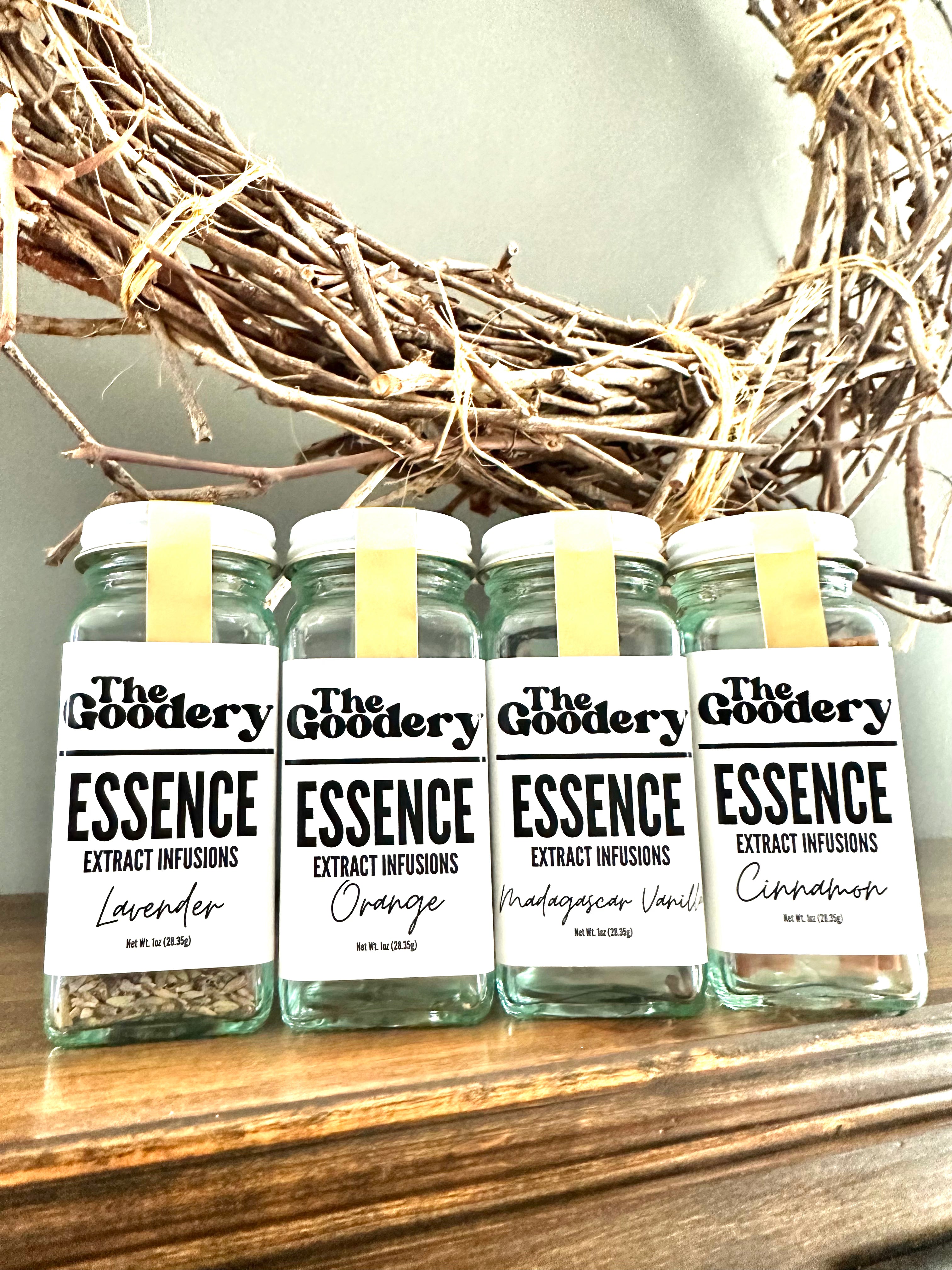 The Goodery Essence Extract Infusions in glass jars, featuring Lavender, Orange, Madagascar Vanilla, and Cinnamon flavors, displayed on a wooden shelf with a rustic wreath in the background.