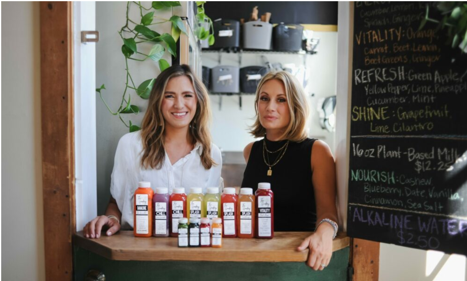 The Goodery founders smiling behind juice bar counter with fresh cold-pressed juices and wellness shots on display