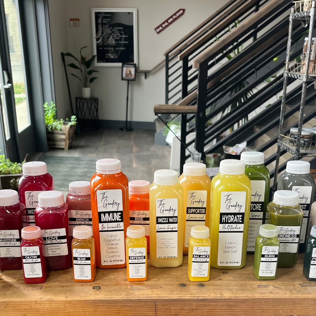 The Goodery juice collection displayed on a wooden table in Stillwater, featuring colorful bottles of cold-pressed juices including Immune, Hydrate, Supporter, and Swizzle Water.
