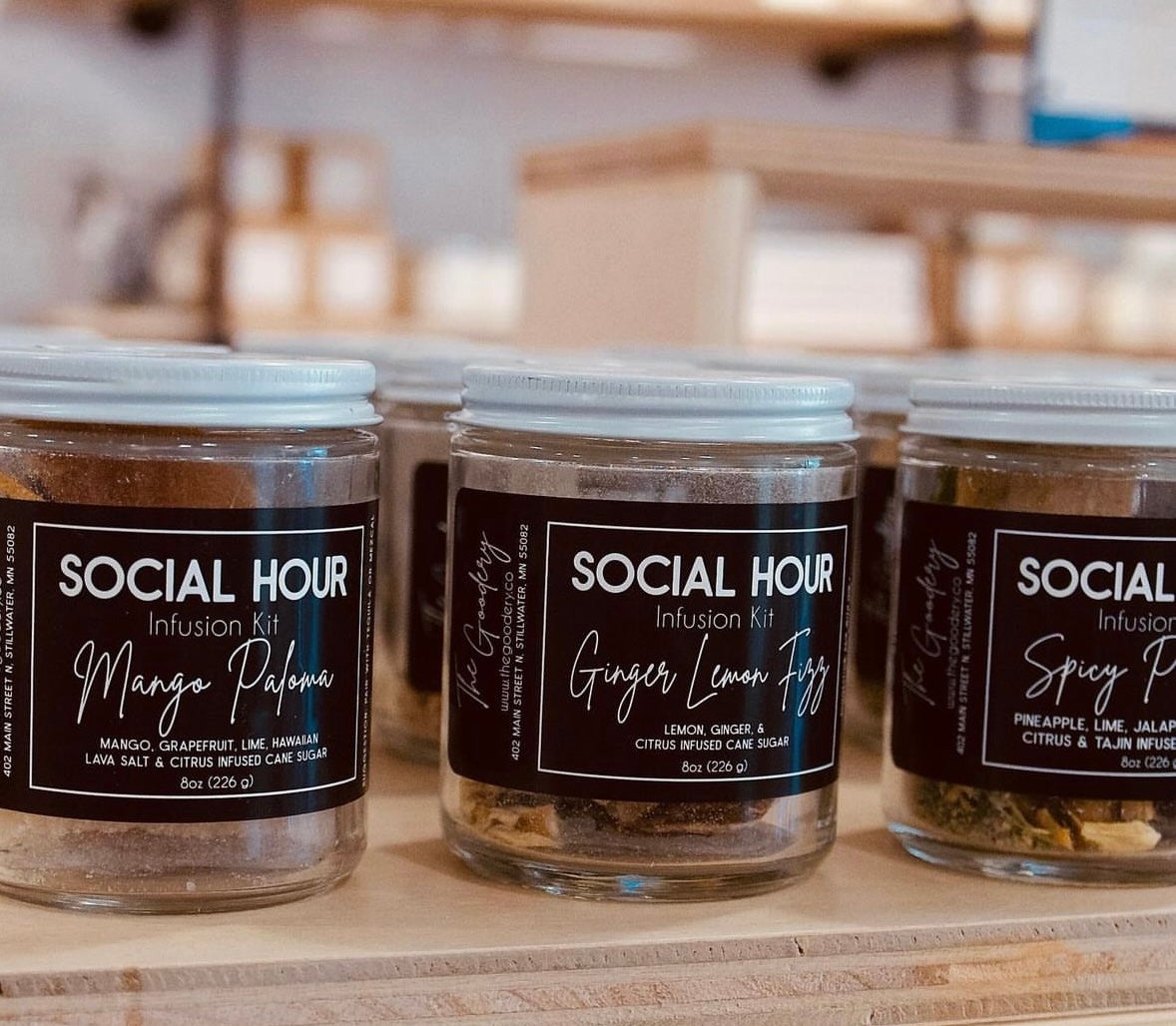 The Goodery Social Hour cocktail and mocktail infusion kits in glass jars with flavors Mango Paloma, Ginger Lemon Fizz, and Spicy Pineapple displayed on a wooden shelf.