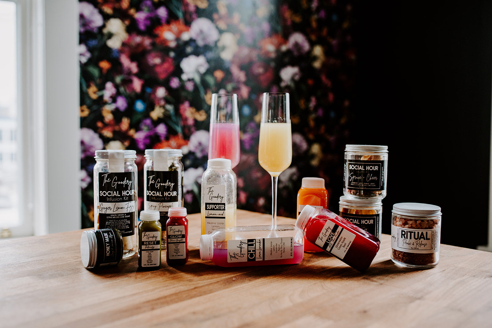 The Goodery Social Hour cocktail infusion kits, juices, and wellness products displayed on a wooden table with floral backdrop