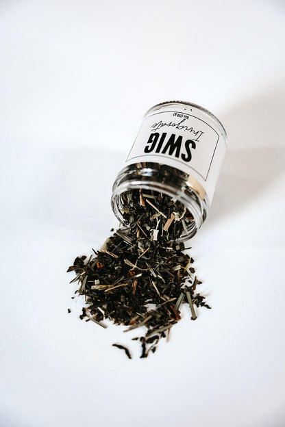 The Goodery Swig Invigorate loose leaf tea jar tipped over with loose tea leaves spilling onto a white background.