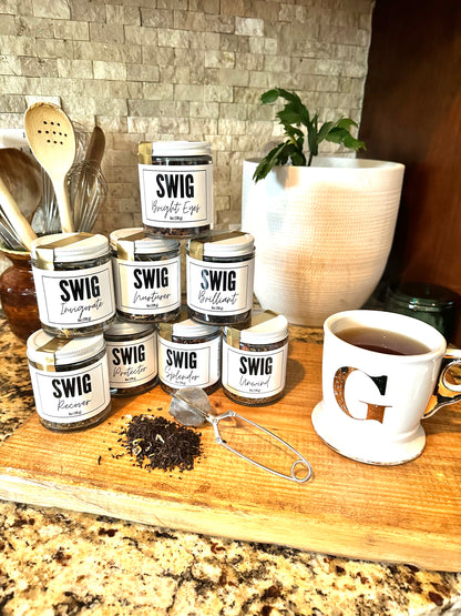The Goodery Swig loose leaf tea collection displayed on a wooden counter with multiple jar flavors stacked beside a cup of tea and tea infuser.