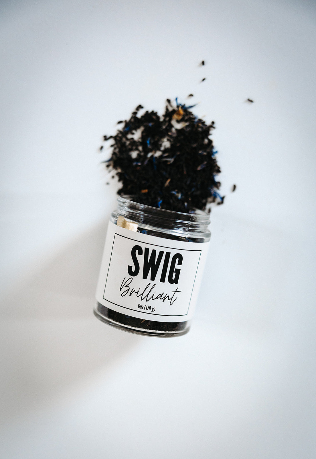 Swig Tea Blend Brilliant loose leaf tea spilling from glass jar with white lid on white background.