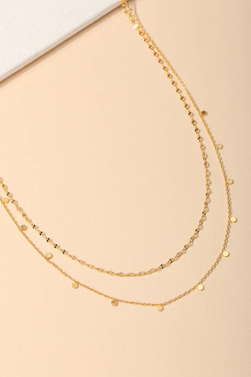 Two layered gold necklaces with small round pendants displayed on a beige background