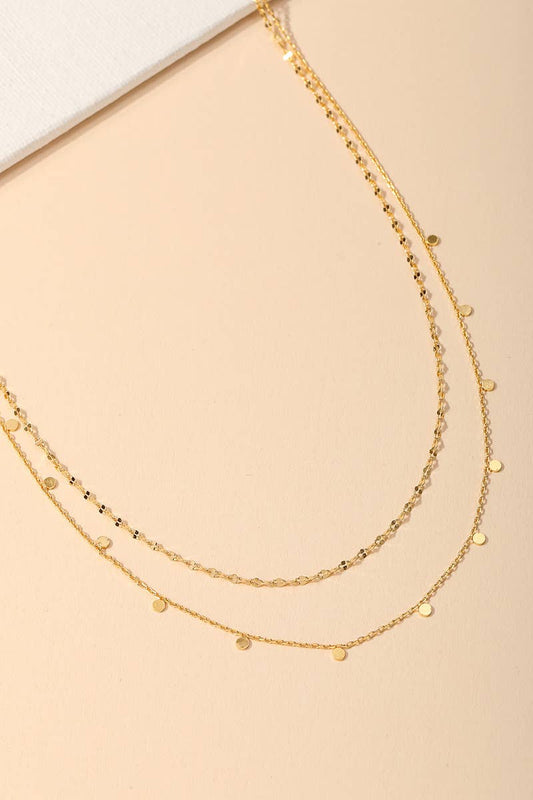 Two layered gold necklaces with small round pendants displayed on a beige background
