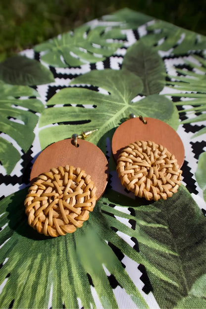 Pair of round wooden earrings with woven straw detail, displayed on leafy fabric background