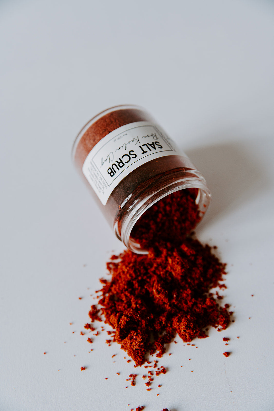 The Goodery Salt Polish skincare product in a glass jar, tipped over with red exfoliating powder spilled onto a white background, natural vegan skincare polish.