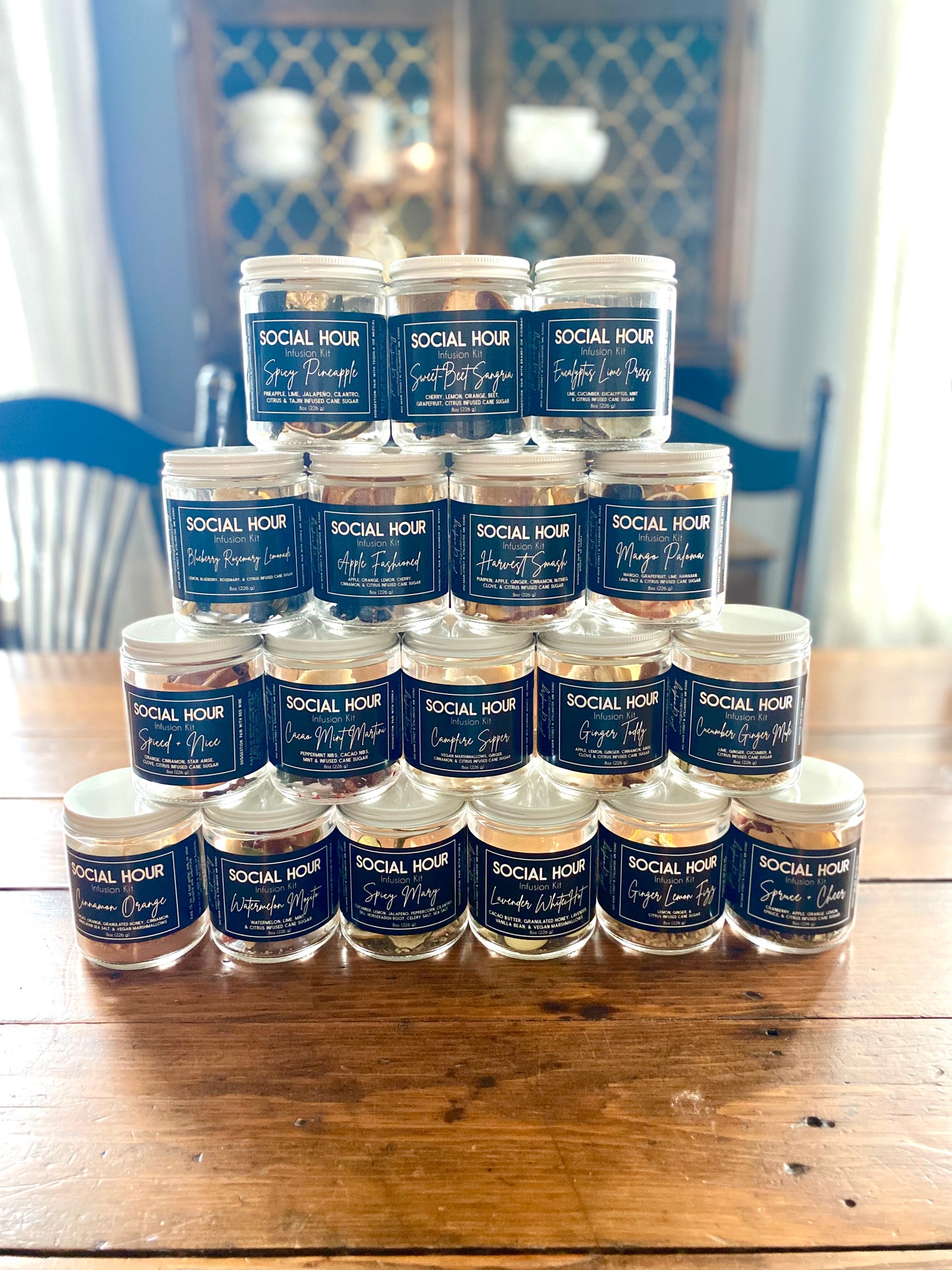 The Goodery Social Hour dried cocktail infusion kit collection in glass jars stacked in a pyramid display on a wooden table, featuring multiple cocktail infusion flavors.