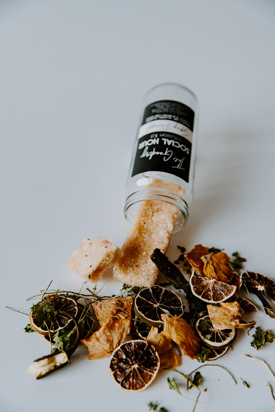 The Goodery Social Hour Infusion Kit in Spicy Pineapple flavor, glass jar tipped over with dried pineapple, citrus, herbs, and sugar cubes spilling onto a white background.